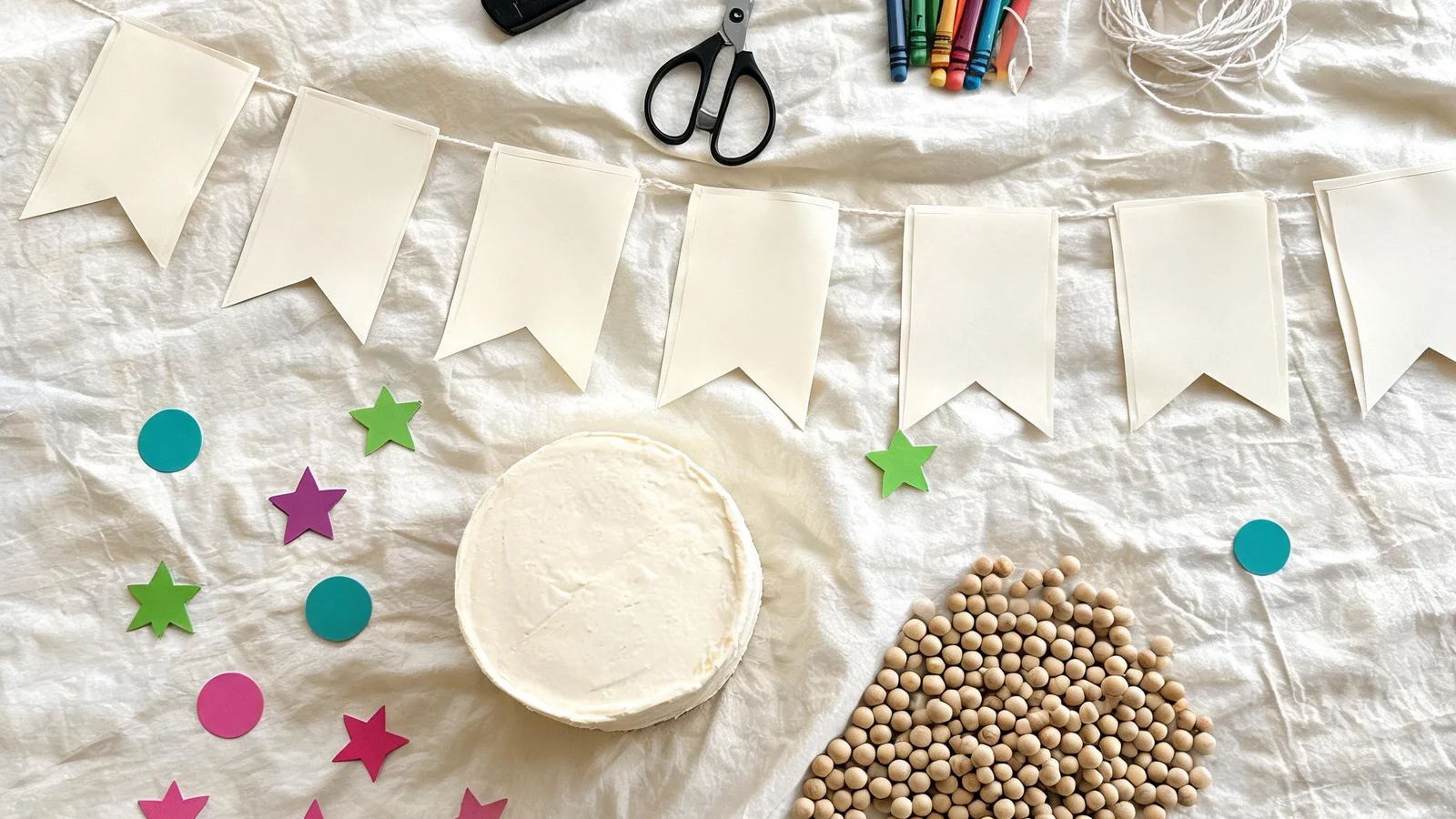 Cream cloth with blank cardstock flags on string, a black punch, scissors, crayons, a plain cake, wooden beads, and paper stars. Illustrative image.