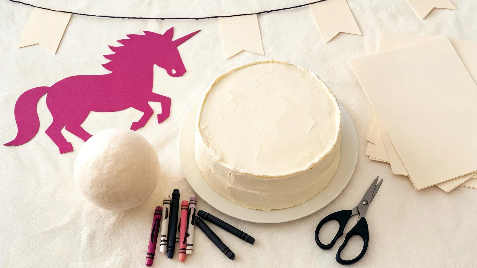 Cream table with a magenta paper unicorn cut-out, blank cardstock flags, a plain cake, a soft ball, crayons, scissors, and blank cards. Illustrative image.