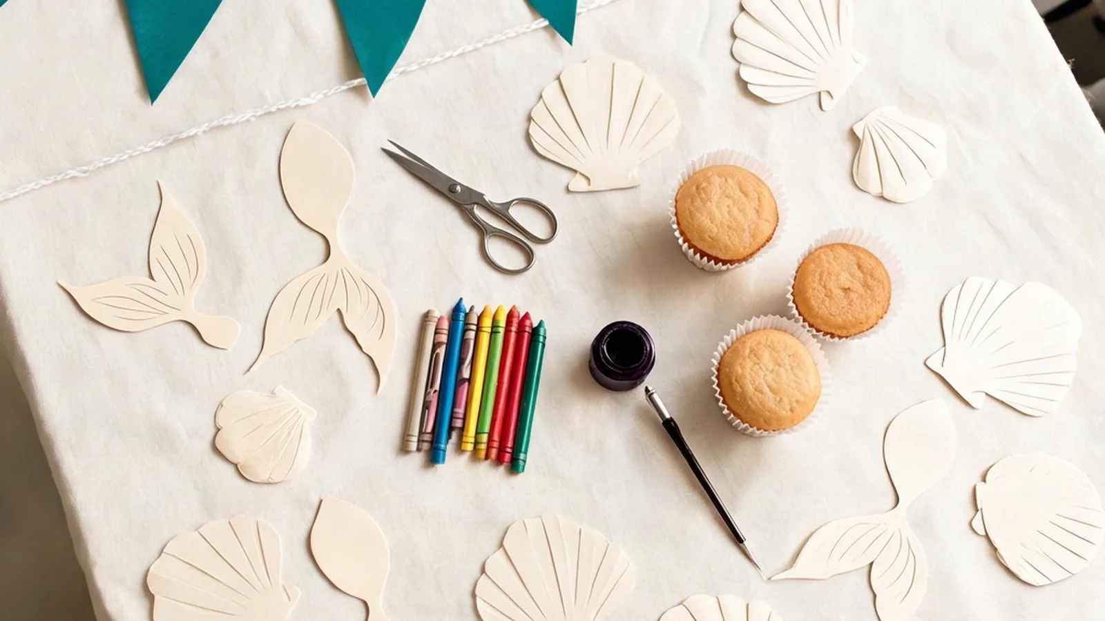 Cream paper mermaid-tail and shell cut-outs, teal cardstock flags on a string, scissors, crayons, and plain cupcakes on a cream table. Illustrative image.