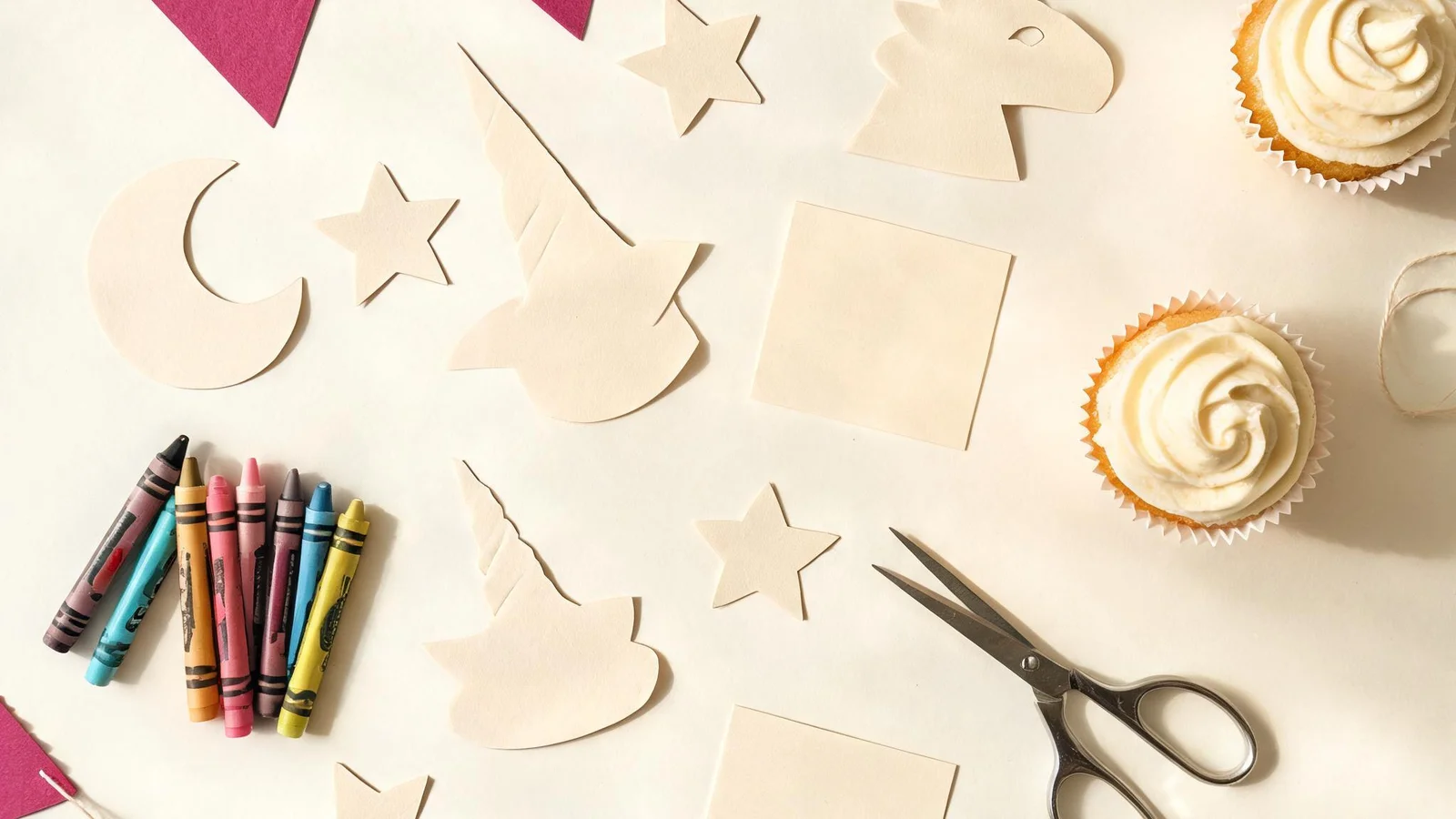 Cream paper unicorn and star cut-outs, magenta flags on string, blank cream cards, scissors, crayons, and frosted cupcakes on a cream table. Illustrative image.