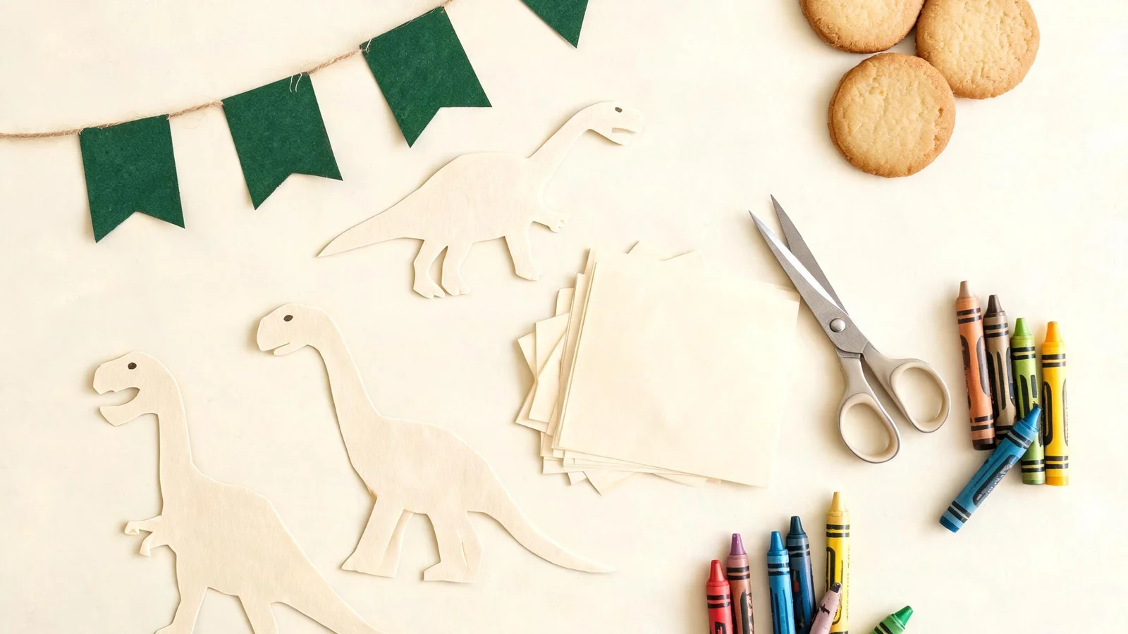 Cream paper dinosaur cut-outs, green flags on string, a stack of blank cream cards, scissors, crayons, and plain cookies on a cream table. Illustrative image.
