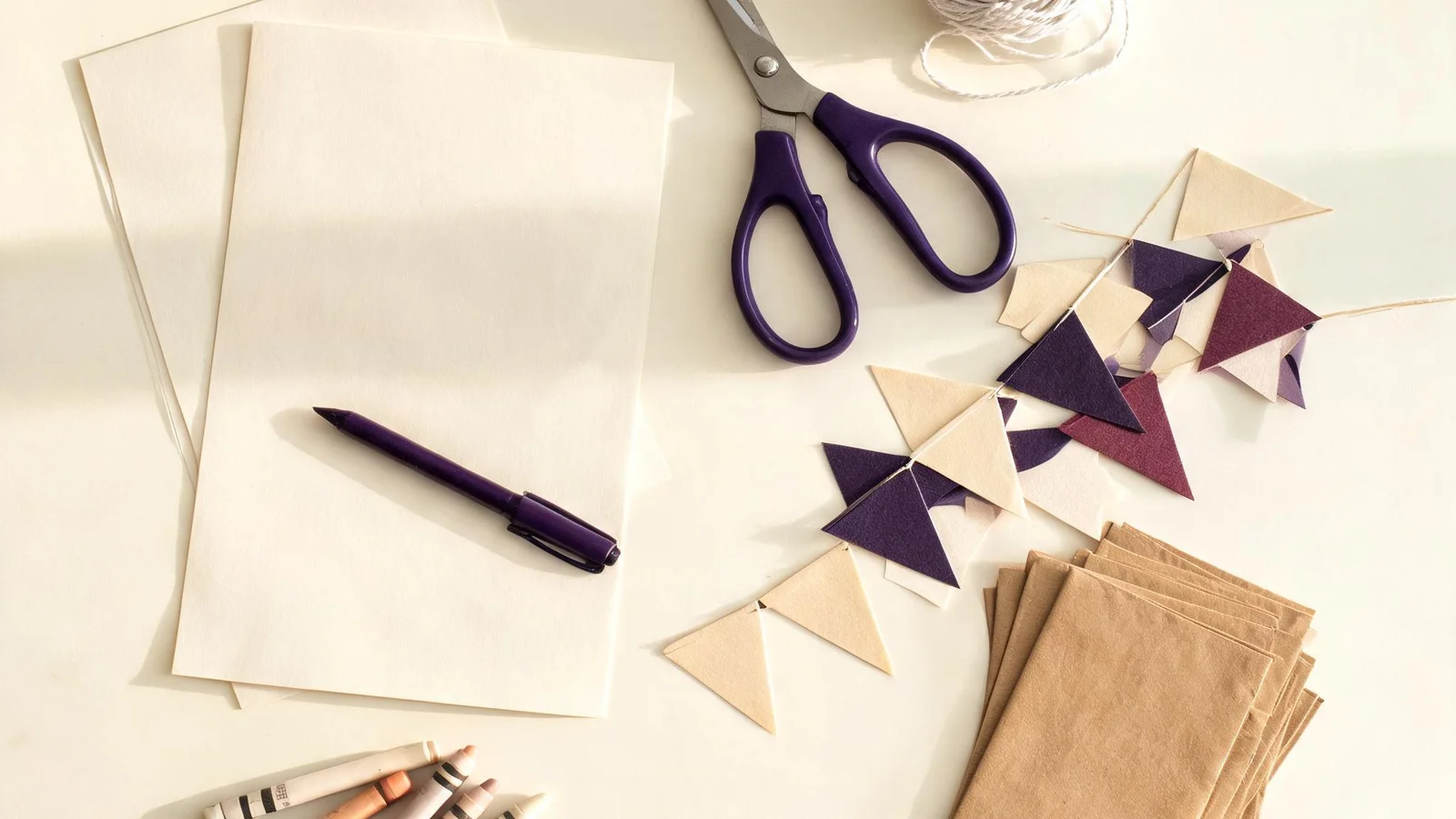 Blank cream sheets, a purple pen, scissors, string, paper triangle flags, crayons, and kraft envelopes on a cream table. Illustrative image.