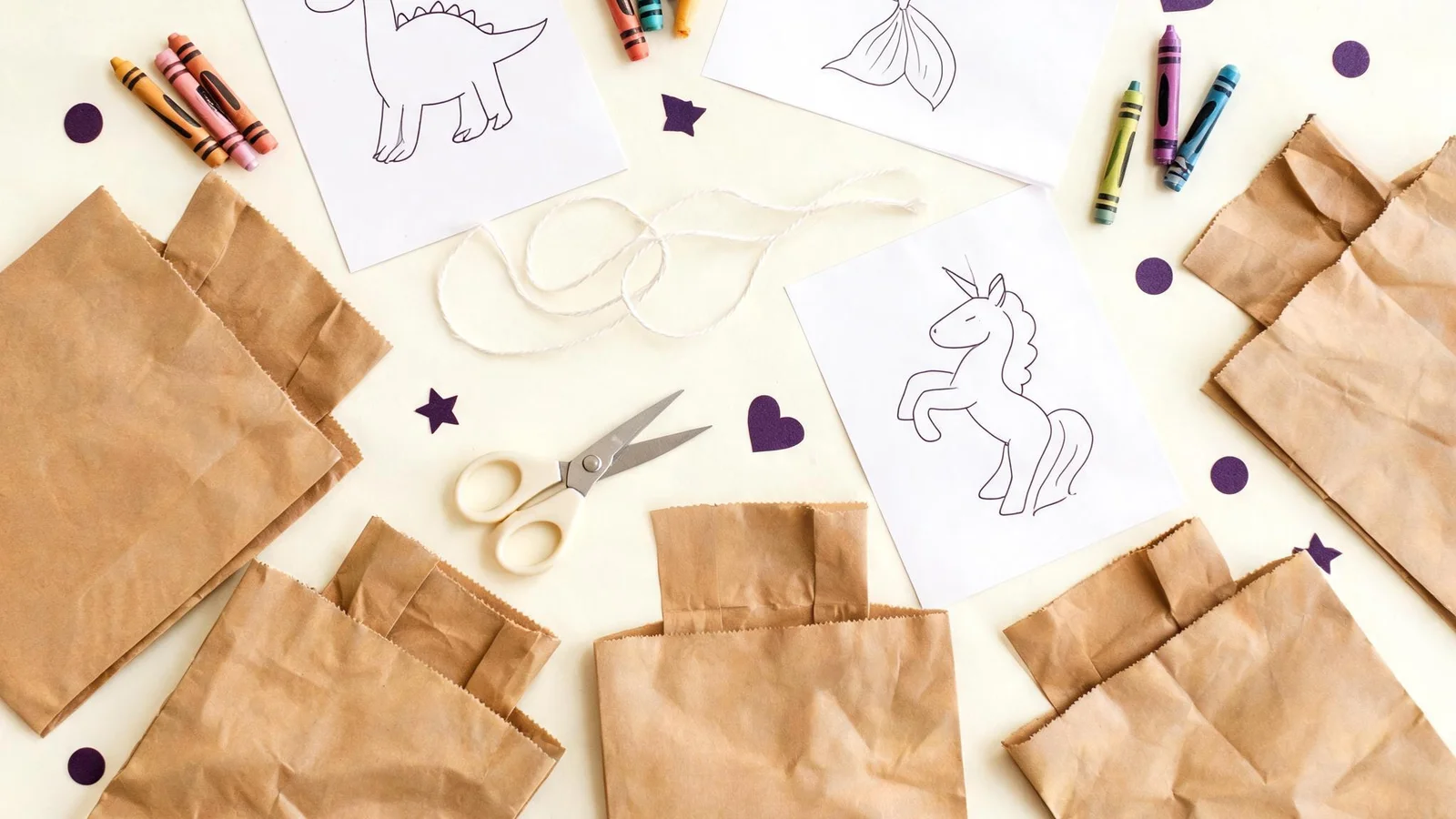 Kraft paper bags, blank line drawings of a dinosaur, a mermaid tail, and a unicorn, crayons, scissors, and string on a cream table. Illustrative image.