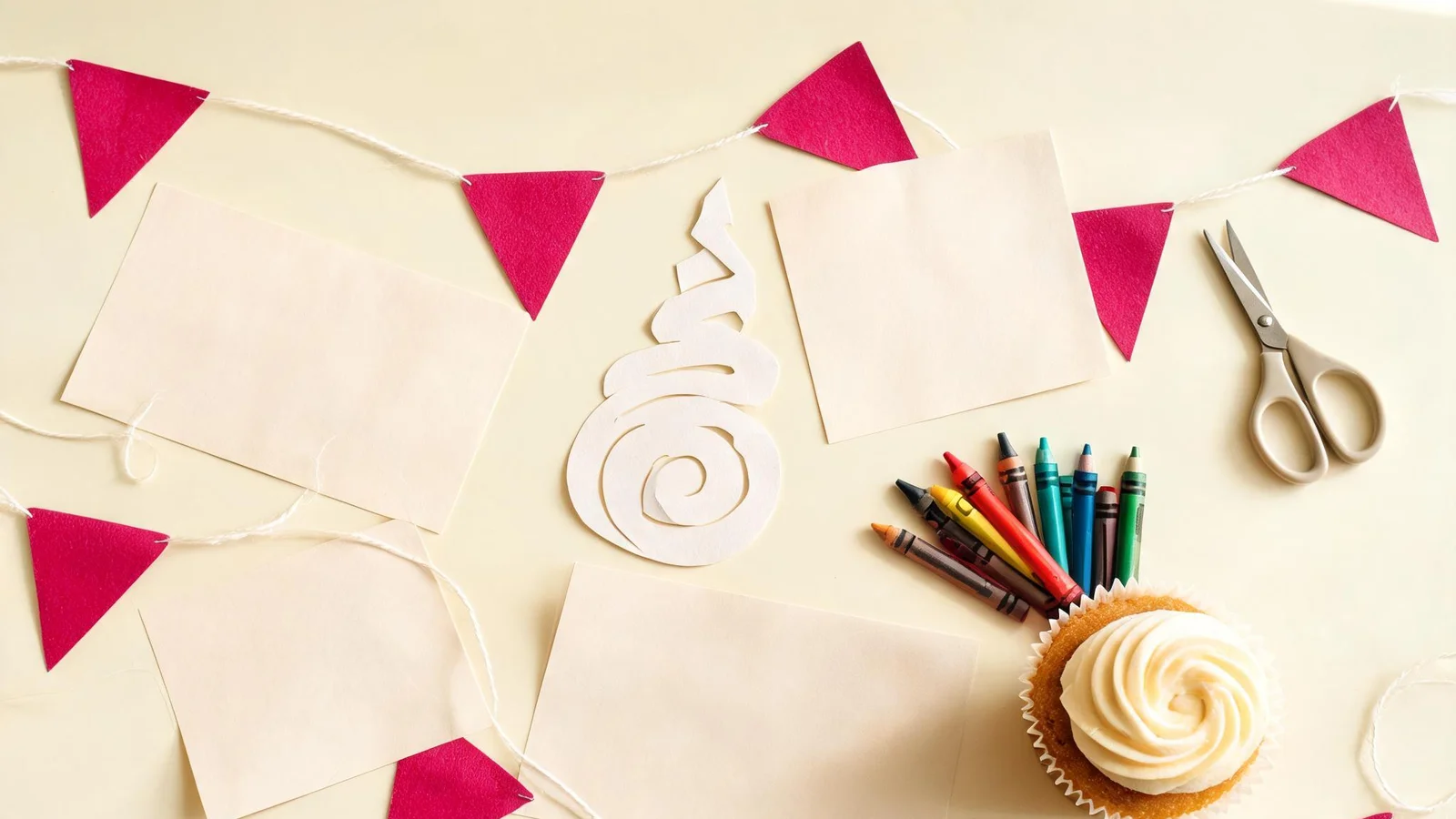 Blank cream cards, magenta paper flags on string, a paper horn cut-out, scissors, crayons, and a cupcake on a cream table. Illustrative image.