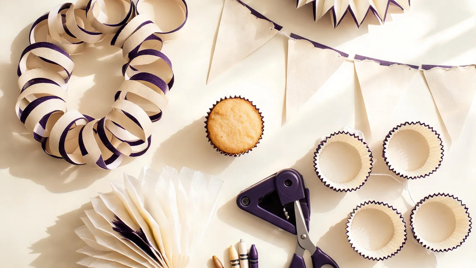 Cream paper chains, triangular flags on string, a paper fan, cupcake liners, scissors, a hole punch, and crayons on a cream table. Illustrative image.