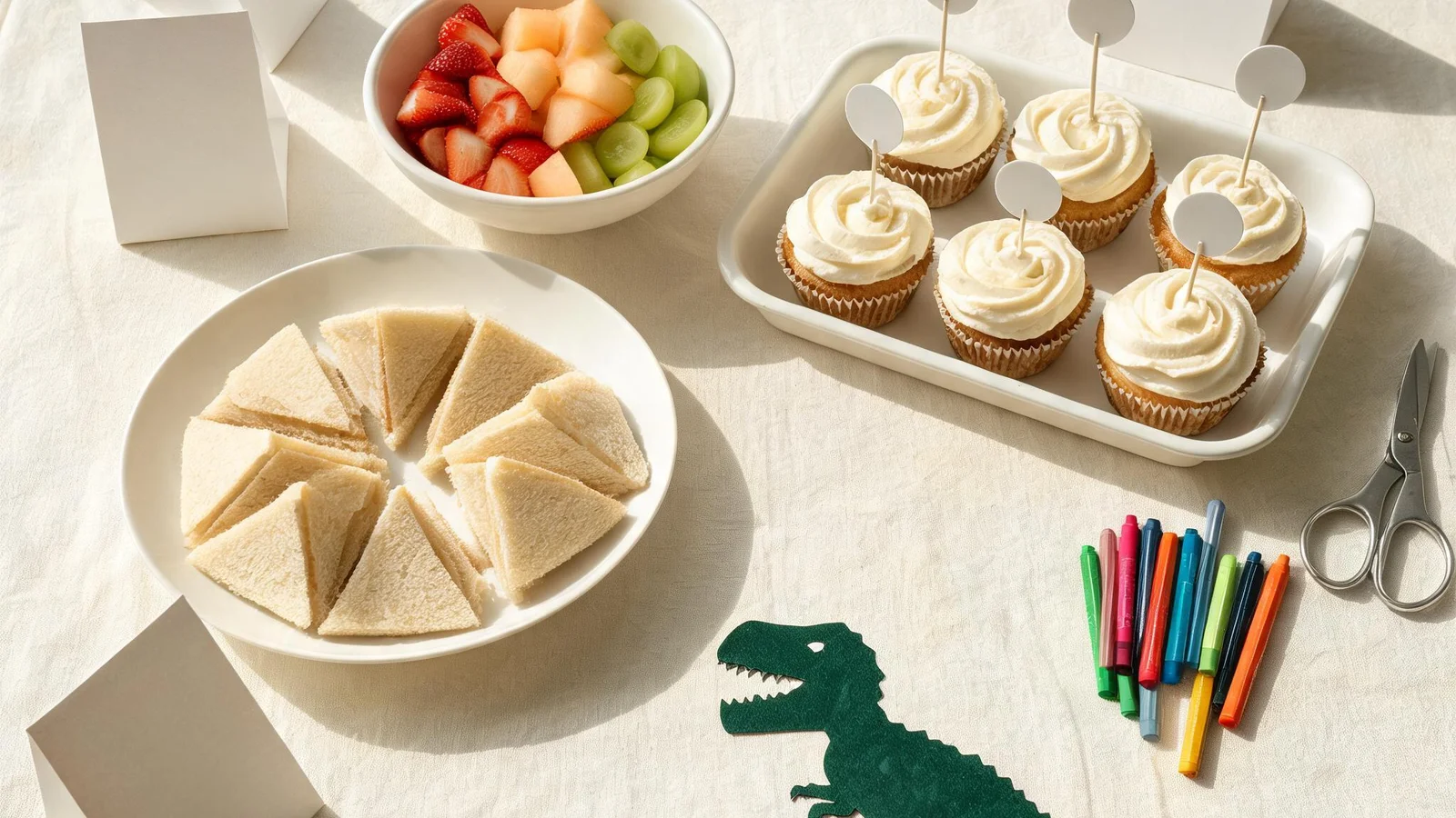 Sandwich triangles, a bowl of strawberries, melon, and grapes, frosted cupcakes with blank paper circles on picks, folded blank cards, a green dinosaur cut-out, markers, and scissors on a cream cloth. Illustrative image.