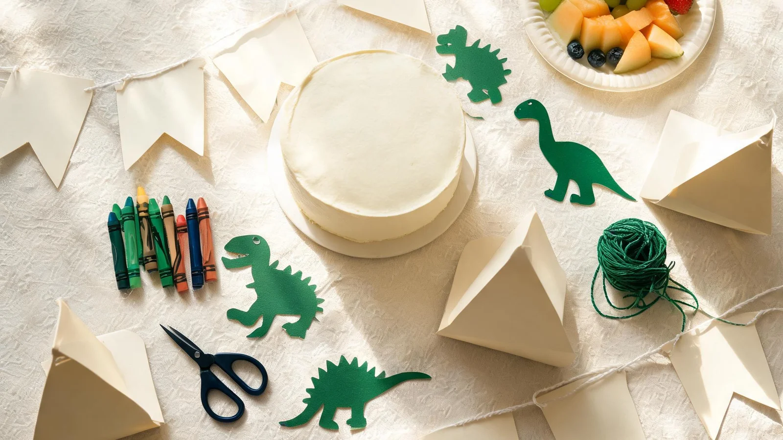 Cream table with a plain cake, green paper dinosaur cut-outs, blank cardstock flags on string, folded paper tents, crayons, scissors, and a plate of fruit. Illustrative image.