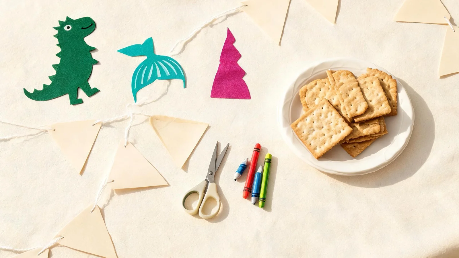 Green paper dinosaur, teal paper mermaid tail, and magenta paper horn cut-outs on a cream table with blank flags, string, scissors, crayons, and crackers. Illustrative image.