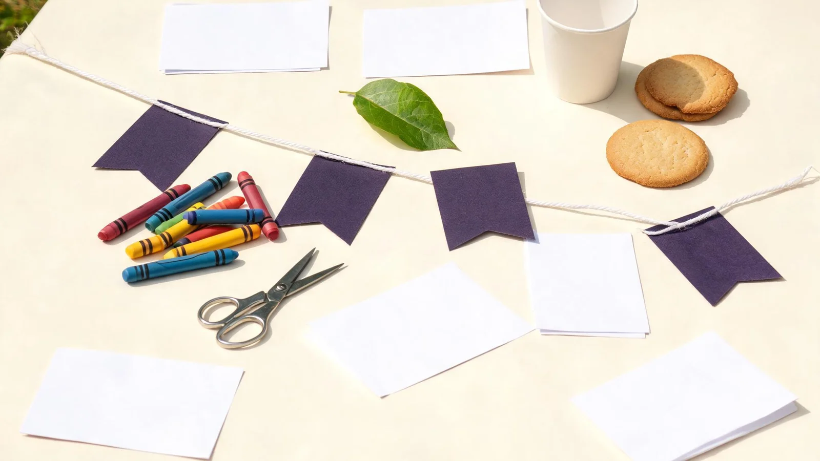 Ink-purple cardstock flags on white string, blank paper cards, crayons, scissors, a green leaf, a paper cup, and cookies on a cream table outdoors. Illustrative image.