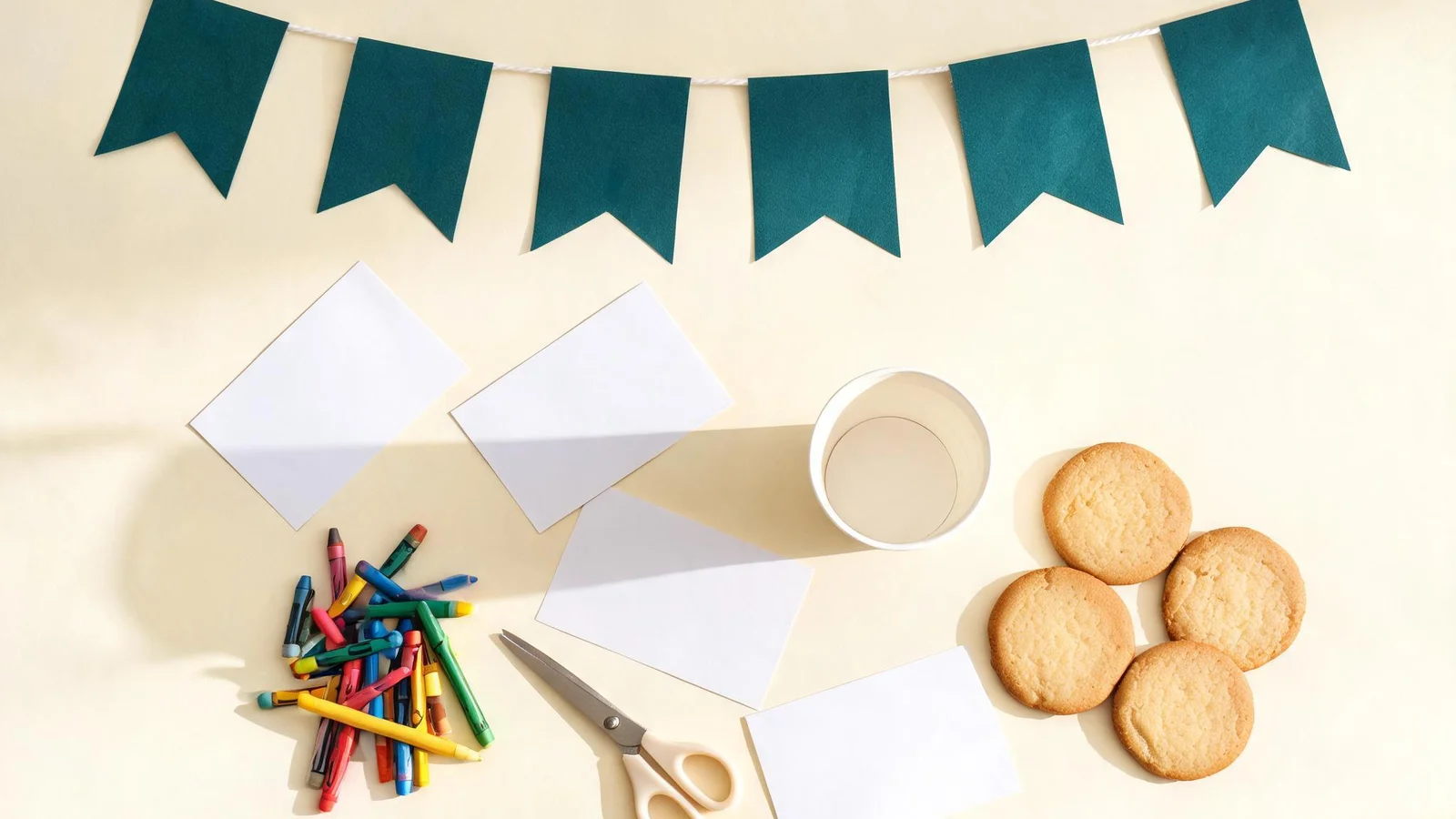 Teal cardstock flags on white string, blank paper cards, crayons, scissors, a paper cup, and cookies on a cream table. Illustrative image.