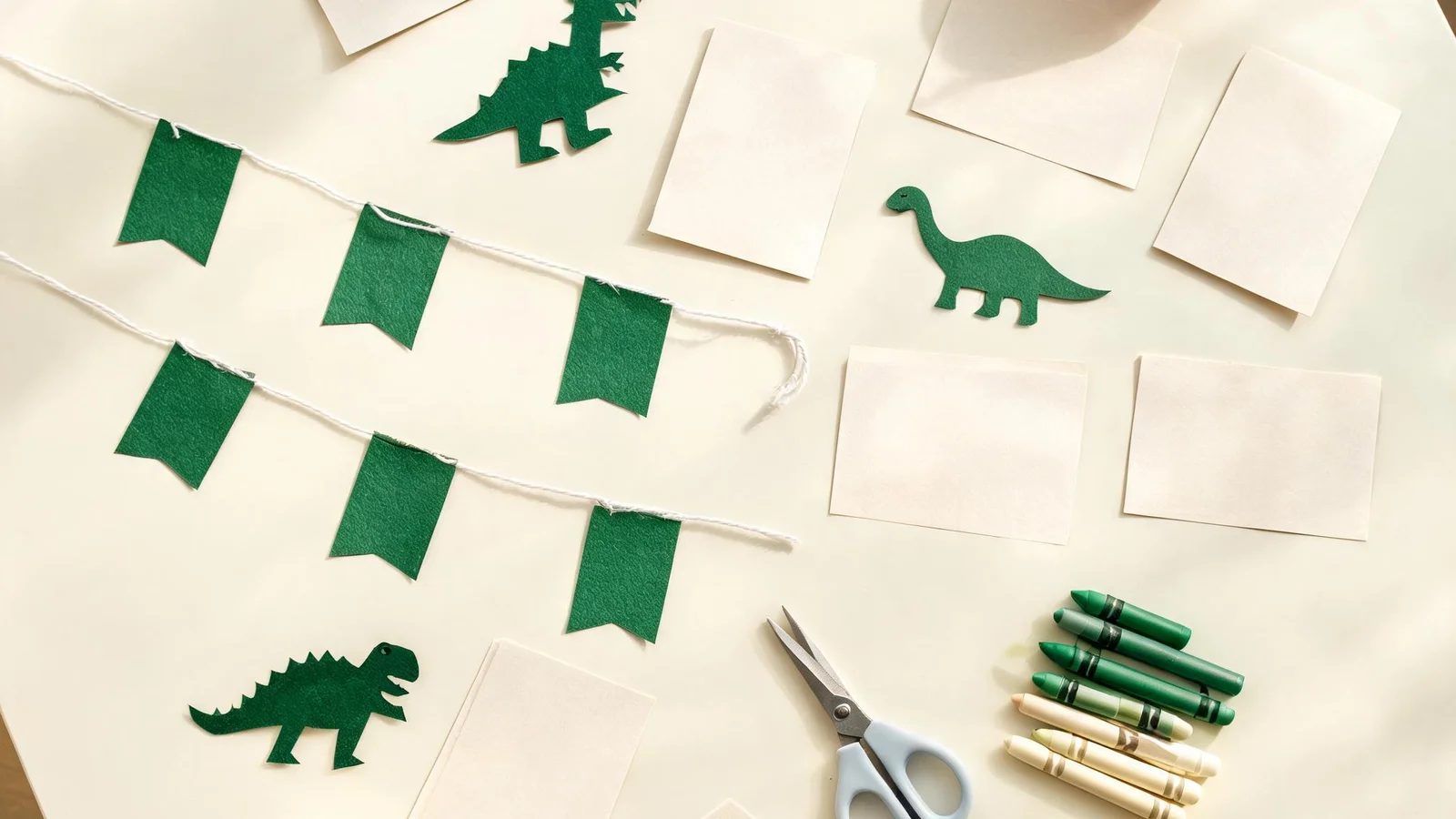 Blank cream cards, green paper dinosaur cut-outs, green flags on string, scissors, and crayons on a cream table. Illustrative image.